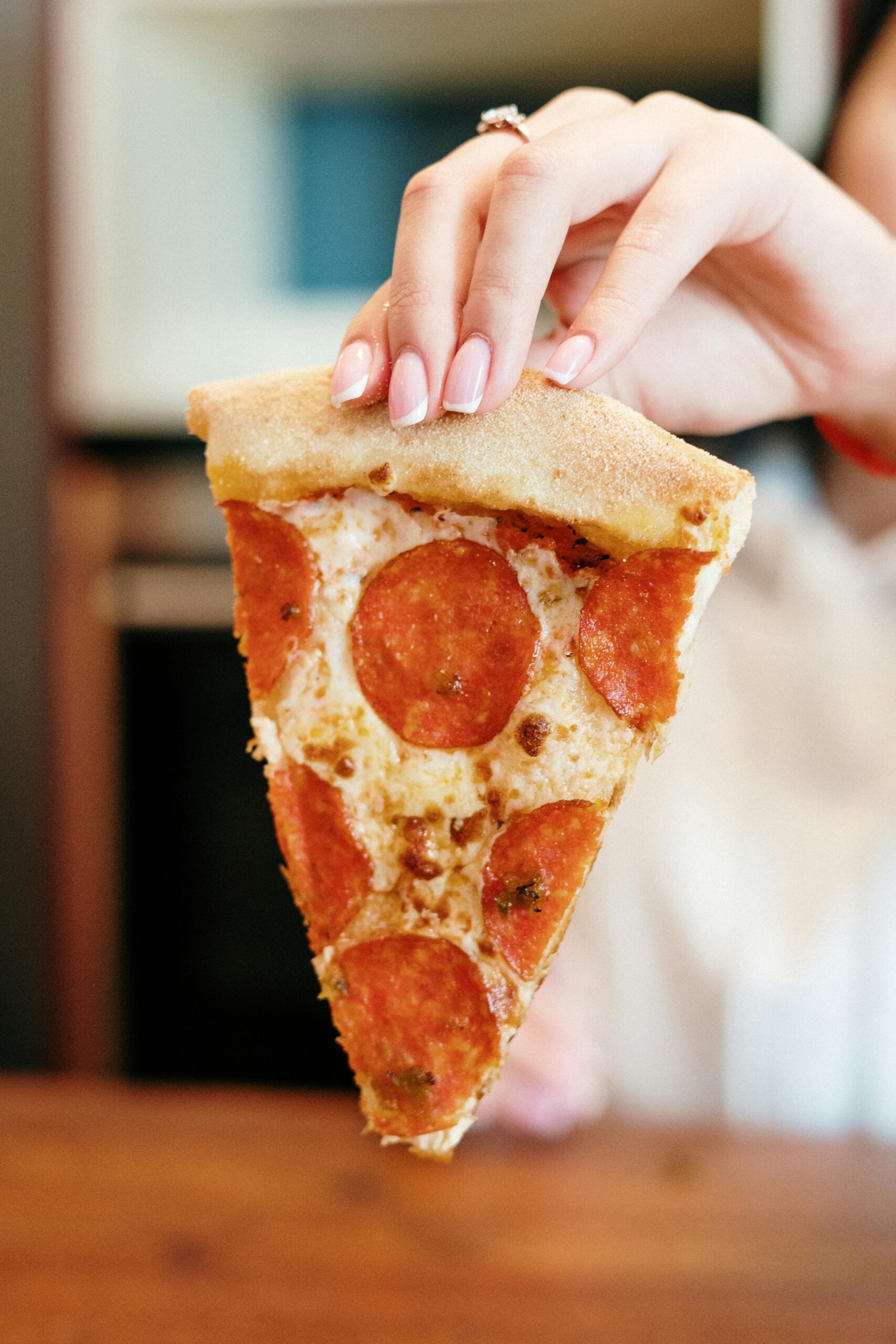 A hand with a polished manicure holds a pepperoni pizza slice indoors.