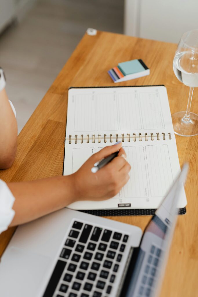 Individual writing in a planner at a desk, accompanied by a laptop and a glass of water.