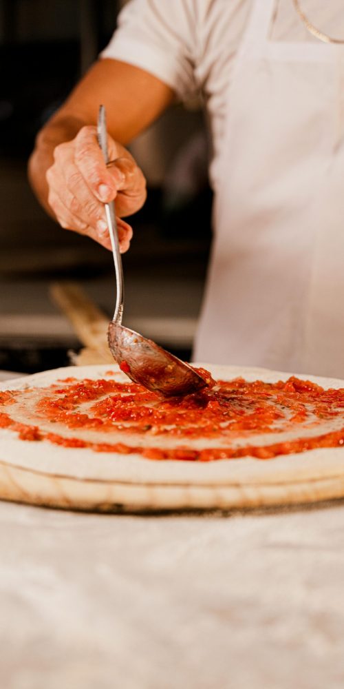 Chef spreading tomato sauce on pizza dough in a kitchen setting with a spoon.